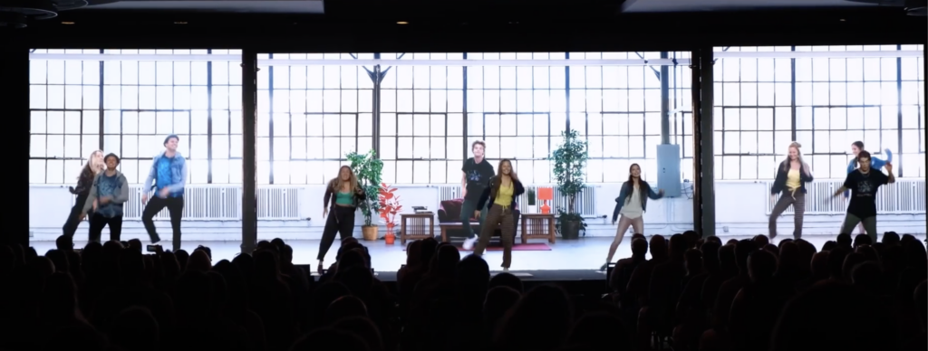 Five people are energetically dancing on a stage in front of a large window backdrop. The backdrop shows couches, plants, and a bright, open space. The audience is visible in silhouette.